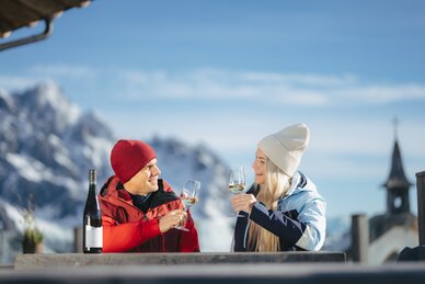 Ein Mann und eine Frau stoßen mit Weißwein an, im Hintergrund verschneite Berge und Kirchturm | © Ski amadé