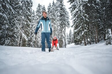 A man pulls a laughing woman on a sled along a snowy forest path. | © Ski amadé