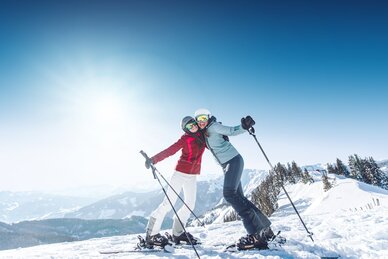 Zwei Frauen posieren mit Skistöcken im Schnee bei Sonnenschein und Bergpanorama in Ski amadé