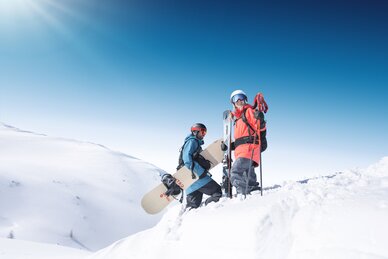 Zwei Wintersportler stehen mit Ski und Snowboard auf einer verschneiten Bergkuppe