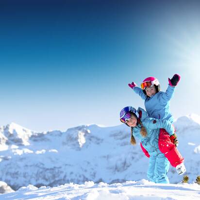 Zwei Kinder in bunter Skikleidung spielen lachend im Schnee vor verschneiten Bergen