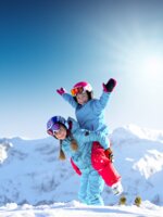 Zwei Kinder in bunter Skikleidung spielen lachend im Schnee vor verschneiten Bergen