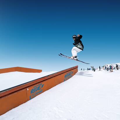 Skifahrer springt im Snowpark über Rail-Element, im Hintergrund warten weitere Wintersportler