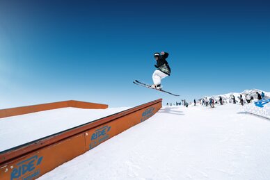 Skifahrer springt im Snowpark über Rail-Element, im Hintergrund warten weitere Wintersportler