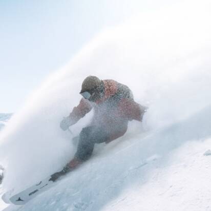 Snowboarder rides down steep slope in deep powder snow, spray clouds in the sunlight
