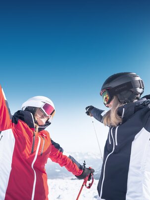 Zwei Skifahrerinnen stehen sich mit ausgestreckten Armen gegenüber, fröhlich im Schnee