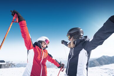Zwei Skifahrerinnen stehen sich mit ausgestreckten Armen gegenüber, fröhlich im Schnee