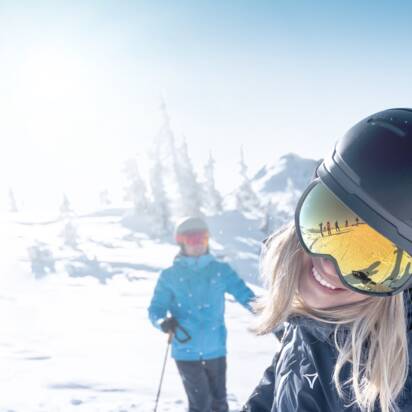 Frau mit Helm und Skibrille lacht in die Kamera, im Hintergrund zwei Skifahrer im Schnee