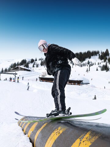 Junge Skifahrerin balanciert auf gelb-schwarzem Rail im Snowpark bei Sonnenschein
