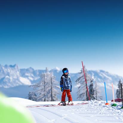 Kind mit Helm und Weste steht auf sonniger Übungspiste mit Skiern vor verschneiten Bergen
