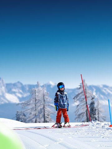 Child wearing helmet and vest stands on sunny training slope with skis in front of snowy mountains