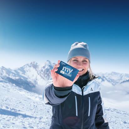 Frau in Skijacke hält my Ski amadé Friends Card vor schneebedeckten Alpen bei strahlendem Sonnenschein