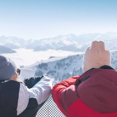 Zwei Personen in Winterkleidung stehen auf Terrasse und schauen auf verschneite Berge mit Skiern im Vordergrund