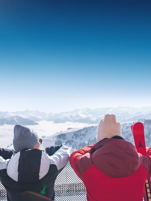 Two people in winter clothes stand on terrace overlooking snowy mountains with skis in the foreground