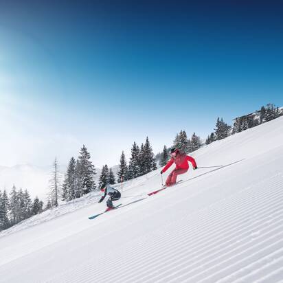 Zwei Skifahrer fahren parallel auf frisch präparierter Skipiste mit Pulverschnee und Sonnenlicht in Ski amadé