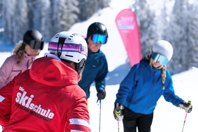 Skilehrerin in roter Jacke unterrichtet Gruppe von Skifahrern mit Helm und Brille auf sonniger Piste in Ski amadé
