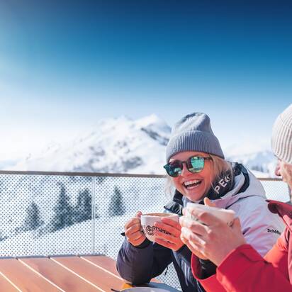 Zwei Personen in Skijacken trinken heiße Getränke auf Terrasse mit Blick auf verschneite Berge bei Sonnenschein