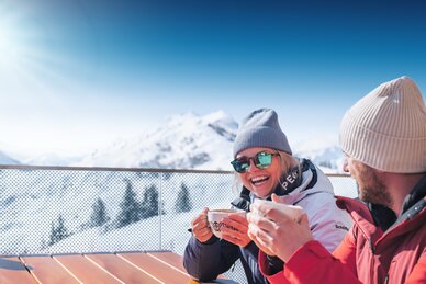 Zwei Personen in Skijacken trinken heiße Getränke auf Terrasse mit Blick auf verschneite Berge bei Sonnenschein