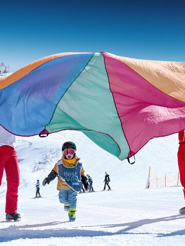 Children in Best Learn2Ski vests play under colourful parachute on snowy slope with ski instructors in the background