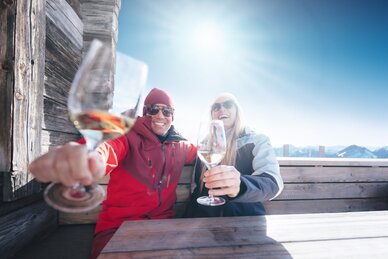 Zwei Skifahrer stoßen mit Weißwein auf Holzbank vor Hütte in Ski amadé an, Sonne und Berge im Hintergrund