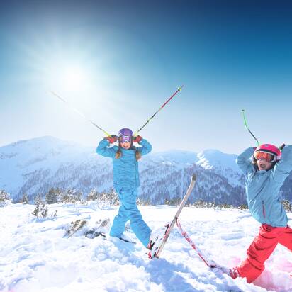 Zwei Kinder stehen im Tiefschnee und halten sich die Skistöcke an den Helm, so dass sie aussehen wie Hörner, und haben den Mund weit offen