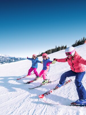 Drei Kinder halten sich an den Händen und fahren nebeneinander die Piste hinunter wobei sich die Skispitzen der einzelnen Kinder berühren und sie lachen dabei