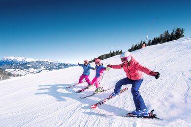Drei Kinder halten sich an den Händen und fahren nebeneinander die Piste hinunter wobei sich die Skispitzen der einzelnen Kinder berühren und sie lachen dabei