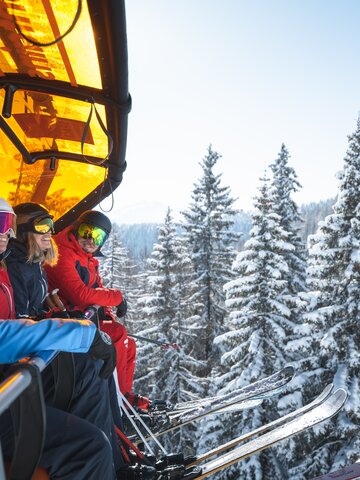 Vier Skifahrer mit Helmen und Skibrillen sitzen auf einem Sessellift mit orangener Haube über verschneiten Tannen. | © Ski amadé