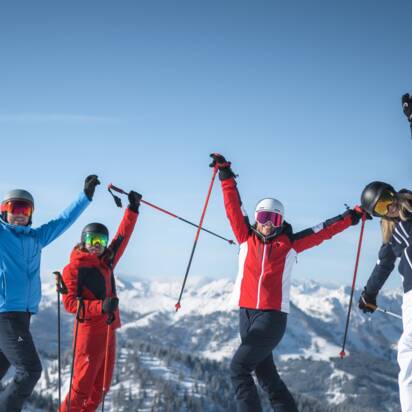 Four friends in ski equipment stand on the piste and stretch their arms joyfully in the air. | © Ski amadé