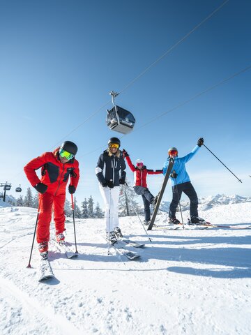 Skifahrer freuen sich und stehen auf der Piste und über ihnen schwebt eine Gondel im strahlend blauen Himmel. | © Ski amadé