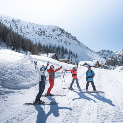 Vier Wintersportler stehen jubelnd vor Almhütten in verschneitem Tal, umgeben von Bergen, Sonne und blauen Himmel.