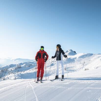 Zwei Skifahrer stehen nebeneinander auf frisch präparierter Skipiste mit Sonne, Sessellift und Alpenblick