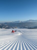 Zwei Personen fahren mit den Skiern eine frisch präparierte Piste hinunter und wirbeln den Schnee auf