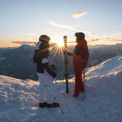 Zwei Skifahrer in Winterkleidung stehen mit Skiern auf schneebedecktem Gipfel bei Sonnenaufgang in den Bergen