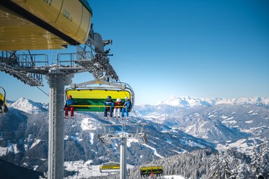 Skifahrer in bunten Skianzügen fahren mit dem Sessellift, Alpenpanorama mit Schneebergen im Hintergrund.