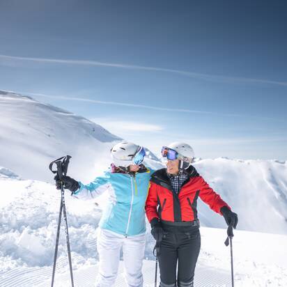 Zwei Skifahrerinnen (Best Ager) umarmen sich lachend auf der Piste, vor verschneiten Bergen mit Skistöcken.