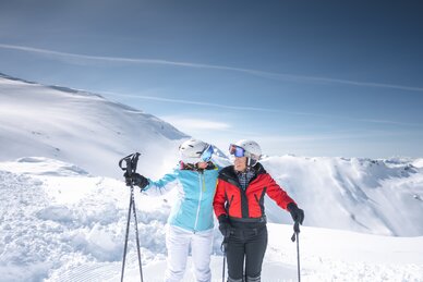 Zwei Skifahrerinnen (Best Ager) umarmen sich lachend auf der Piste, vor verschneiten Bergen mit Skistöcken.