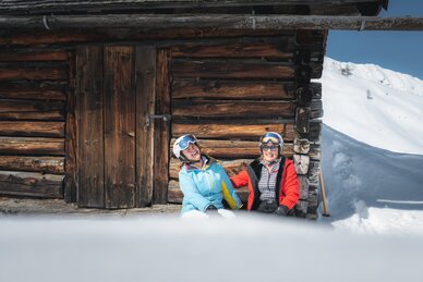 Zwei Skifahrerinnen (Best Ager) lachen vor uriger Holzhütte im Schnee, genießen Sonne und Alpenblick.