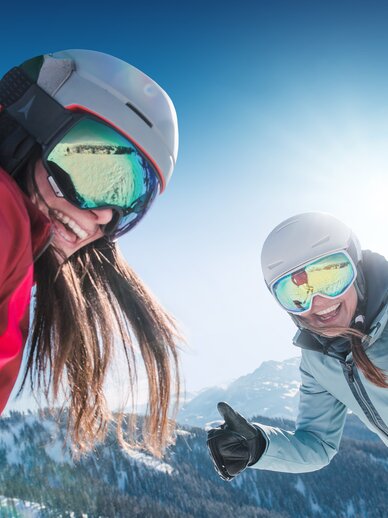 Zwei Skifahrerinnen lachen mit Helm und Skibrille, genießen die Sonne und verschneite Alpen, Pisten im Hintergrund.
