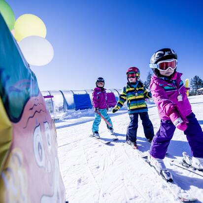 Three kids in colorful ski suits ski on a sunny slope between ski school features, smiling and motivated.