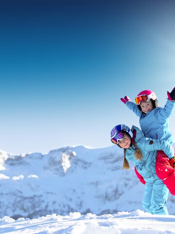 Zwei Mädchen in blauer Skibekleidung spielen lachend im Schnee vor verschneiten Bergen bei Sonnenschein, eine hat Hände hoch.